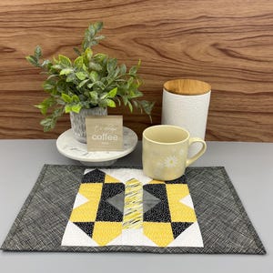 May include: A black and white quilted placemat with a yellow and grey geometric design. A white mug with a floral pattern sits on the placemat. A small wooden sign with the text "It's always coffee time" sits on a white marble coaster.