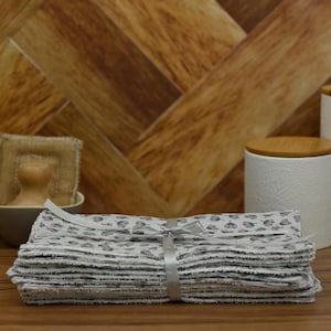 May include: A stack of grey and white patterned reusable cloths tied with a white ribbon. A small potted plant, a bowl with a wooden handled brush, and two white ceramic canisters with wooden lids are in the background.
