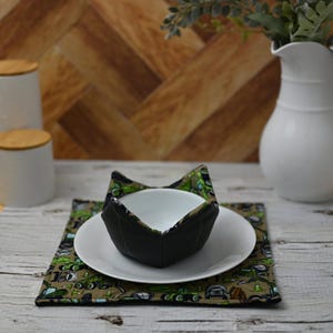May include: A black and green fabric bowl cosy with a white ceramic bowl inside. The bowl cosy is sitting on a white plate and a green and brown placemat with a pattern of construction vehicles.