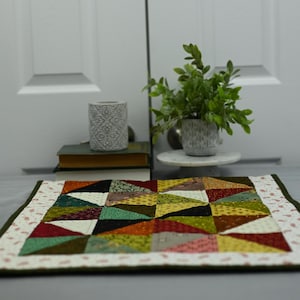 May include: A small, colourful patchwork quilt with a green border. The quilt is made up of various shades of brown, green, red, and yellow triangles. The quilt is sitting on a grey surface in front of a white door.