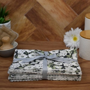 May include: A bundle of floral napkins, patterned with white flowers and green leaves on a black background, tied with a grey ribbon. Other items include a small bowl with a wooden scrubber and a white flower.