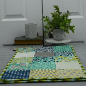 May include: A patchwork table runner featuring a green, blue and white colour scheme. The runner is made from squares of different fabrics and has a green border.