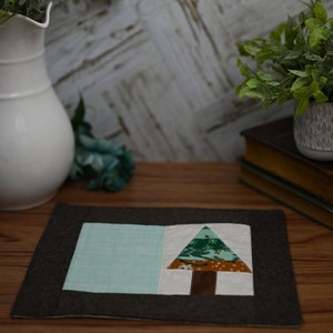 May include: A handmade, rectangular placemat with a dark grey border. The centre features a patchwork design with a light blue square and a white square with a green and brown Christmas tree. The placemat is on a wooden surface.