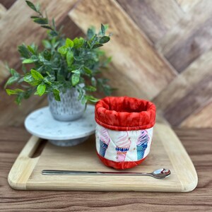 Peut inclure: Un étui à cônes de crème glacée en tissu rouge et blanc avec une cuillère assortie repose sur une planche à découper en bois. Une petite plante dans un pot blanc est posée sur une assiette blanche à gauche de l'étui. L'étui présente un motif répété de cônes de crème glacée.