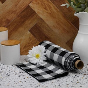 May include: A roll of black and white buffalo check fabric with a white daisy on top. The fabric is rolled up and tied with a brown ribbon. The fabric is on a table with a white ceramic jar and a white ceramic jug.