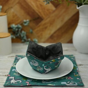 May include: A teal blue fabric bowl cosy with a black interior and a whimsical dragon print. The cosy is sitting on a white plate and a teal blue placemat with a dragon print.