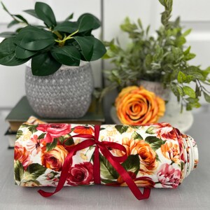 May include: A white fabric with a floral print of red, orange, and pink roses. The fabric is rolled up and tied with a red ribbon.