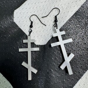May include: A pair of silver-colored earrings featuring a stylized Orthodox cross design. The earrings have a hook closure and a small black bead at the top. The crosses have a three-bar design and a slanted lower bar.