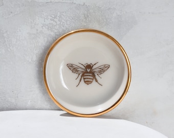 May include: A small white ceramic trinket dish with a gold rim and a gold bee design. The bee has a heart in its body.