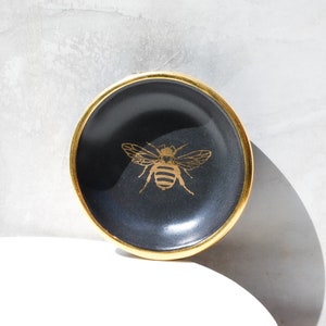 May include: A small, round, black dish with a gold rim and a gold bee design in the center. The bee is detailed with wings and body segments. The dish is likely ceramic or porcelain.