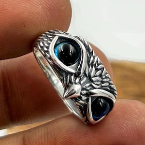 Peut inclure: Bague argentée représentant un hibou. Les yeux du hibou sont noirs avec un contour bleu. La bague présente une surface texturée et détaillée, avec les plumes et le bec du hibou clairement définis. Un bijou unique.