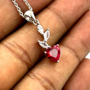 Puede incluir: Un collar de plata con un colgante de piedra roja en forma de corazón. El colgante está adornado con piedras claras en forma de hoja y cuelga de una delicada cadena. La joya se muestra sobre un fondo neutro.