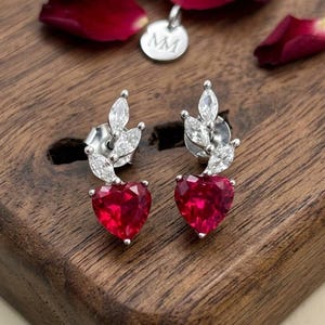 May include: A pair of silver-toned stud earrings featuring a heart-shaped red gemstone at the base and three marquise-cut clear stones arranged in a leaf-like pattern above. The earrings include a small circular silver tag engraved with the letters MM.