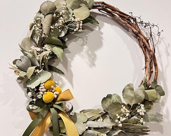 May include: A decorative wreath made of dried eucalyptus leaves, small white flowers, and yellow pom-poms. The wreath is adorned with a mustard yellow and olive green ribbon. The base is made of intertwined brown branches.