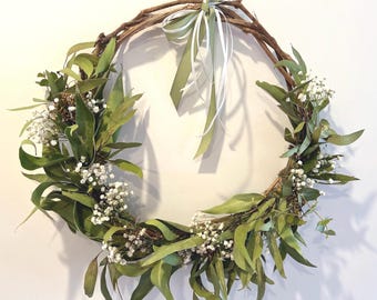 May include: A circular wreath made of interwoven brown branches, adorned with green leaves and small white flowers. A green and white ribbon is tied at the top for hanging. The wreath is a natural, rustic decoration.
