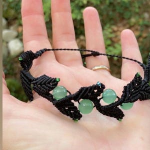 Può includere: Un braccialetto in macramè nero caratterizzato da una serie di motivi annodati a forma di foglia. Il design incorpora perline rotonde, lisce e traslucide di colore verde chiaro intervallate tra i segmenti di corda scura, con minuscole perline.