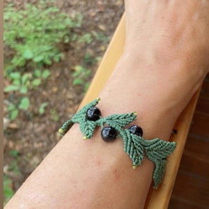 Può includere: Un braccialetto in macramè realizzato con cordino verde salvia intrecciato in una serie di motivi a forma di foglia. Il design è arricchito da tre perline nere rotonde e lisce posizionate tra le foglie e piccole perline dorate sulle punte.