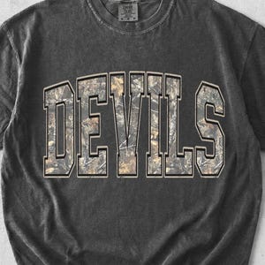 May include: A charcoal grey short-sleeve t-shirt with a washed-out finish. The front features the word DEVILS in large, block-style letters with a camouflage pattern fill in shades of brown, tan, and green, outlined in a thin light-colored border.