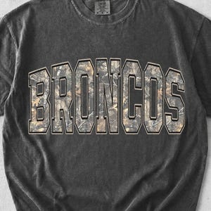 Puede incluir: Una camiseta de manga corta gris carbón con un acabado lavado vintage. La parte delantera presenta la palabra BRONCOS en letras grandes de estilo bloque con un relleno de patrón de camuflaje marrón y verde y un borde negro.