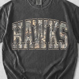 May include: A charcoal grey short-sleeve t-shirt with a crew neckline. The front features the word HAWKS in large, block-style lettering with a camouflage pattern fill in shades of brown, tan, and green, outlined in a thin white border.