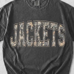 May include: A charcoal grey short-sleeve t-shirt with a crew neckline. The center of the chest features the word JACKETS in large, block-style capital letters. The letters have a camouflage pattern in shades of brown, green, and tan.