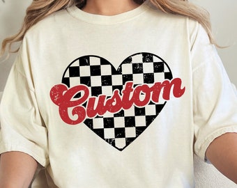 May include: Cream-coloured t-shirt with a black and white chequered heart and the word "Custom" in red script. The shirt has a relaxed fit and is worn with blue jeans. The overall aesthetic is casual and vintage-inspired.