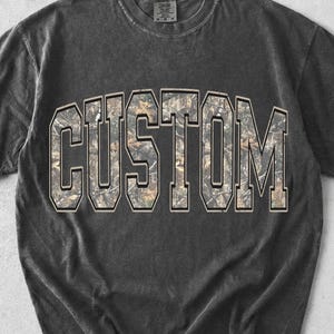 May include: A dark gray t-shirt with the word "CUSTOM" in large, block letters across the chest. The letters are filled with a camouflage pattern. The shirt has a vintage, washed appearance.