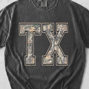 Peut inclure: T-shirt gris foncé avec les lettres "TX" dans un motif camouflage. Les lettres sont soulignées d'une couleur beige clair. Le t-shirt a un aspect vintage et usé.
