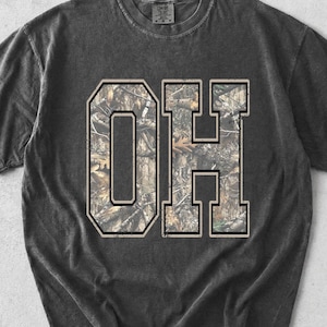 May include: Dark gray t-shirt featuring the letters "OH" in a collegiate style. The letters are filled with a camouflage pattern and outlined in a contrasting color. The shirt has a vintage, worn look.