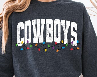 Puede incluir: Sudadera gris oscuro con la palabra "COWBOYS" en letras blancas desgastadas. Debajo de la palabra hay una cadena de luces navideñas de colores. La sudadera es de cuello redondo.