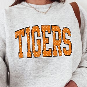 May include: A light gray heathered crewneck sweatshirt featuring the word TIGERS in large, orange, block-style letters with a white polka dot pattern. The sweatshirt has long sleeves with ribbed cuffs and a ribbed neckline, paired with light blue denim jeans.