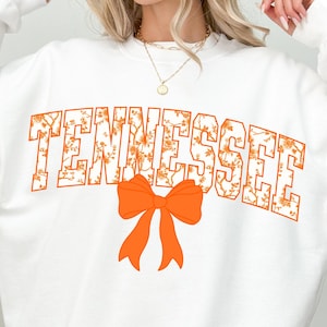 May include: White sweatshirt with the word "TENNESSEE" in orange, floral-patterned lettering. An orange bow graphic is centered below the text. The person is wearing black shorts.