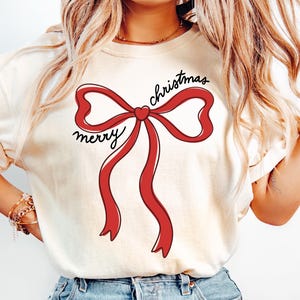 Könnte beinhalten: Cremefarbenes T-Shirt mit einer großen roten Schleife und dem Schriftzug "merry christmas" in schwarzer Schrift. Das Shirt wird mit hellblauen Jeans kombiniert. Der Look hat einen lässigen, festlichen Stil, passend zur Weihnachtszeit.