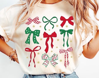 May include: A white t-shirt with a festive design of red and green bows with gold stars. The bows are arranged in a random pattern on the shirt.