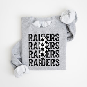 Pode incluir: Camisola cinzenta com um gráfico de raio branco e preto e a palavra "RAIDERS" repetida quatro vezes.