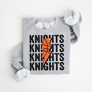 May include: A light gray sweatshirt with the word "KNIGHTS" stacked four times in black, with an orange basketball and lightning bolt design in the center. The sweatshirt has a crew neck and long sleeves.