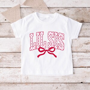May include: White short-sleeved t-shirt with the text "LIL SIS" in red, heart-filled letters. A red bow is centered below the text. The shirt is displayed on a light wood surface with a burlap banner in the background.
