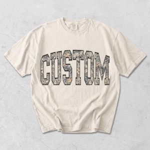 May include: Cream-colored t-shirt with the word "CUSTOM" in a camouflage-style font. The letters are outlined in black, creating a bold contrast against the neutral background. The shirt has a classic crew neck and short sleeves.