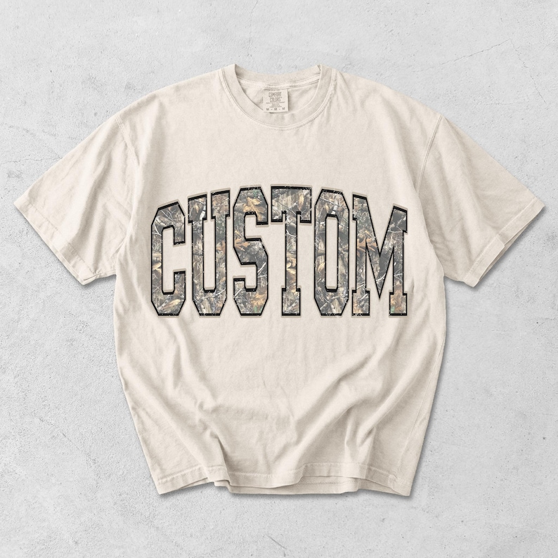 May include: Cream-colored t-shirt with the word "CUSTOM" in a camouflage-style font. The letters are outlined in black, creating a bold contrast against the neutral background. The shirt has a classic crew neck and short sleeves.