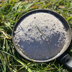 May include: A black metal pan filled with a light gray powder, sitting on a bed of green grass.