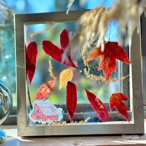 May include: A framed artwork featuring pressed autumn leaves in shades of red, orange, and yellow. A small illustration of a red barn is also included. The frame is a metallic silver color.
