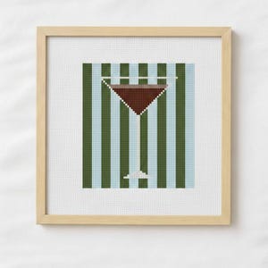 May include: Cross-stitch artwork of a cocktail in a martini glass. The drink is a dark brown color. The background has vertical stripes of blue and green. The artwork is in a light wood frame.