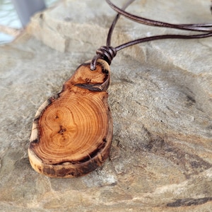 Peut inclure: Un collier pendentif en bois fait à la main. Le pendentif en forme de goutte d'eau révèle le grain naturel du bois dans des tons bruns et beiges. Il est suspendu à un cordon en cuir marron, noué en haut. Le collier repose sur une surface de pierre texturée.