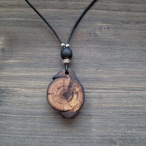 A wooden pendant necklace with a natural, organic design. The pendant features a distinctive shape with visible wood grain and a dark stain. It is suspended from a black cord, with a black bead and silver-toned metal accents. The necklace is shown on a wooden surface.