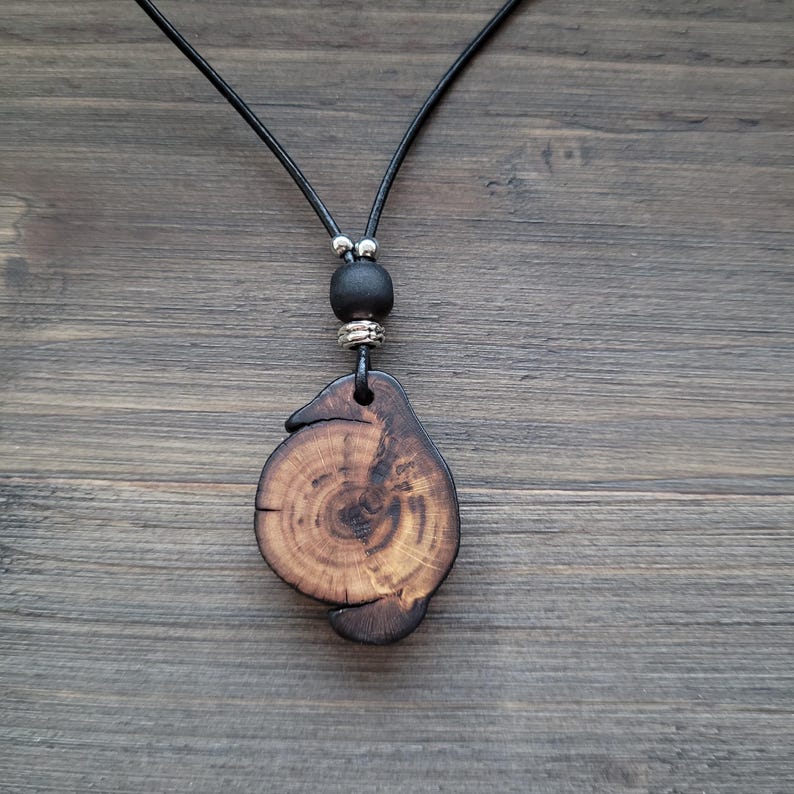 A wooden pendant necklace with a natural, organic design. The pendant features a distinctive shape with visible wood grain and a dark stain. It is suspended from a black cord, with a black bead and silver-toned metal accents. The necklace is shown on a wooden surface.