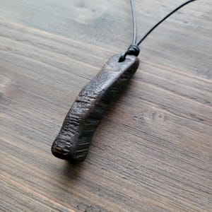 Puede incluir: Un colgante de madera de color marrón oscuro, de forma alargada, con una superficie rugosa y texturizada que presenta surcos y crestas profundos e irregulares. El colgante cuelga de un cordón negro fino que se extiende hacia arriba.