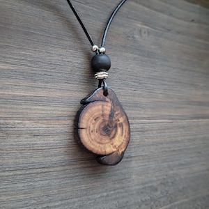 A dark whimsical wooden necklace. A wooden pendant necklace with a natural wood slice pendant. The pendant has visible grain and a dark brown finish. The necklace includes a black leather cord, a black bead, and silver-coloured accents. The pendant is approximately 4 cm in diameter.
