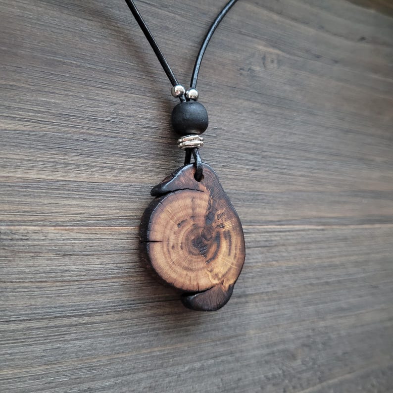 A dark whimsical wooden necklace. A wooden pendant necklace with a natural wood slice pendant. The pendant has visible grain and a dark brown finish. The necklace includes a black leather cord, a black bead, and silver-coloured accents. The pendant is approximately 4 cm in diameter.