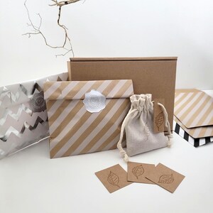 Eco-friendly packaging items. A brown paper bag with diagonal white stripes and a white wax seal, a small drawstring bag, and brown tags and cards with leaf illustrations. A brown cardboard box is in the background.