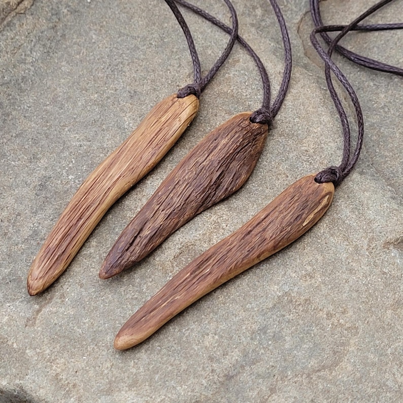 Three handcrafted wooden pendant necklaces made from organic live edge oak wood in long irregular pin shape. The pendant is suspended on a dark brown cotton cord.  The pendants has rich dark live edge wood grooves and natural earthy appearance. These natural oak wood pendants necklaces would be perfect for Pagan, Wicca, Druid or anyone seeking organic, earthy piece of jewelry.